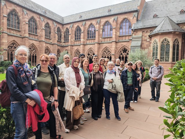 Reisegruppe im Kreuzgang des Mainzer Doms. Die Teilnehmenden stehen vor den historischen roten Sandsteinmauern und gotischen Fenstern.